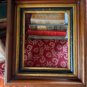 May include: A rectangular wooden picture frame with a dark brown finish, black inner border, and gold accents. The frame surrounds a stack of antique books with red, gray, and cream-colored covers. The background is a red fabric with a gold floral pattern.