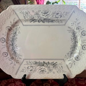 May include: An oval ceramic serving platter in cream with a dark gray floral transferware pattern. The rim features an embossed geometric border, while the center displays detailed illustrations of wildflowers and leaves on all four sides.