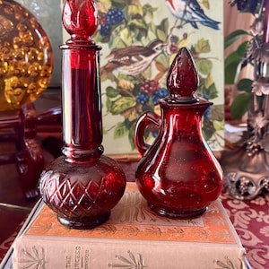 May include: Two antique ruby red glass bottles with stoppers. One is tall and slender with a diamond pattern, the other is a small pitcher shape. They sit on top of vintage books with gold lettering.