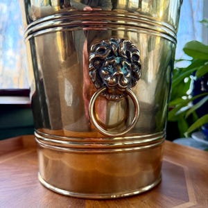 May include: A vintage brass ice bucket with a lion's head ring handle. The bucket has a polished finish and a classic design, perfect for entertaining. The bucket is approximately 8 inches tall and 7 inches in diameter.