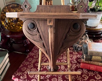 May include: A dark wood shelf with an ornate, carved design. The shelf has a heart-shaped base supported by a bamboo-style stand. The shelf is displayed on a burgundy patterned cloth, with books and decorative items in the background.