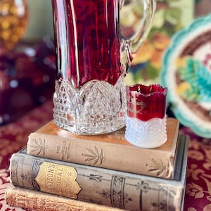May include: A ruby red glass pitcher with a clear glass base and a clear glass cup with a ruby red top. The pitcher and cup are sitting on a stack of three vintage books.