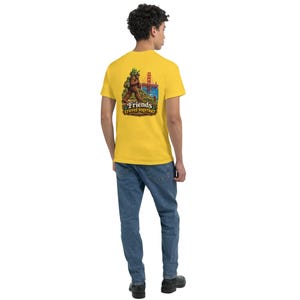May include: A bright yellow short-sleeve t-shirt featuring a graphic on the back. The illustration depicts a brown creature carrying a green alien, with the Golden Gate Bridge in the background and the text Friends travel together below.