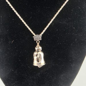 Puede incluir: Un collar de tono plateado con una cadena de cuerda y un colgante en forma de campana. La parte superior del colgante tiene un pequeño dije con forma de casa con la palabra FAMILY grabada. La campana muestra dos figuras estilizadas abrazadas.