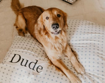 Könnte beinhalten: Ein Golden Retriever liegt auf einem weißen und blauen, gemusterten Hundebett mit dem Namen "Dude" darauf.