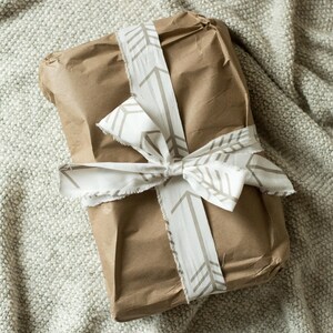 May include: A brown paper gift bag tied with a white ribbon with a brown arrow pattern. The bag is sitting on a light brown textured surface.