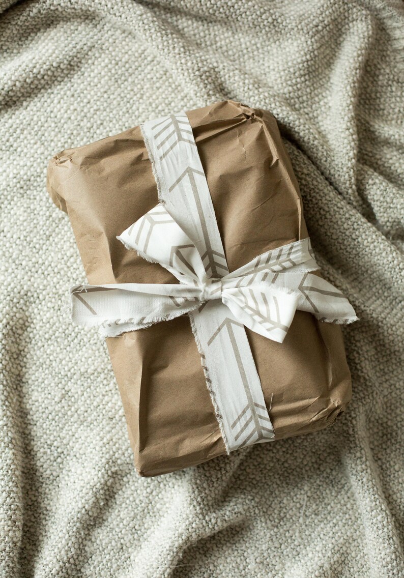 May include: A brown paper gift bag tied with a white ribbon with a brown arrow pattern. The bag is sitting on a light brown textured surface.