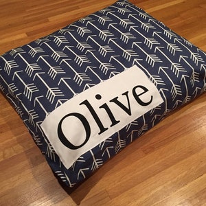 Puede incluir: Una cama para perro azul y blanca con un parche blanco que dice "Olive" en letras negras.