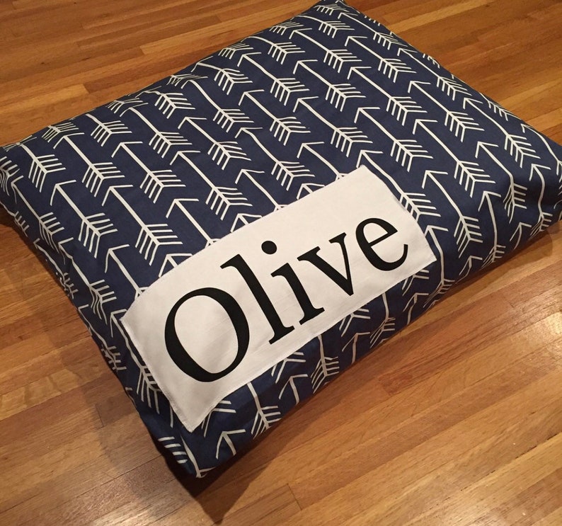 Puede incluir: Una cama para perro azul y blanca con un parche blanco que dice "Olive" en letras negras.