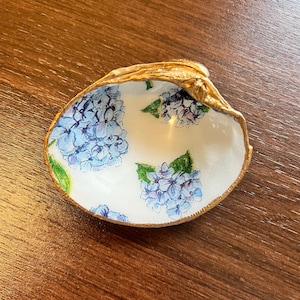 May include: A white seashell dish featuring hand-painted blue hydrangea flower clusters with green leaves on the interior surface. The outer rim and edges of the shell are finished with a metallic gold paint application.