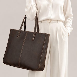 Peut inclure: Un sac cabas en cuir marron foncé présentant une silhouette rectangulaire, deux coutures verticales au centre et deux anses assorties fixées par des rivets métalliques. Le sac présente des surpiqûres contrastées visibles sur les bords.