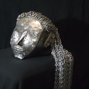 May include: A metallic sculpture of a human head crafted from polished silver-toned metal. The face features defined eyes, a nose, and lips. Long strands of silver-colored metal chains are attached to the top of the head, cascading down the side.