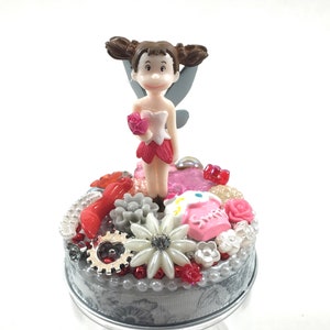 May include: A decorative glass jar with a fairy figurine on top. The fairy is wearing a pink and white dress and holding a pink rose. The jar's lid is adorned with various embellishments, including red and white beads, a gear, and a white flower.