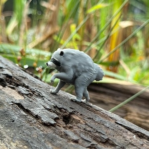 Puede incluir: Una pequeña figura tridimensional de color gris que representa a un mapache de pie sobre sus cuatro patas. El mapache tiene una máscara negra distintiva alrededor de los ojos, un hocico puntiagudo y una postura encorvada. Es de material gris mate.