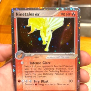 Puede incluir: Una carta coleccionable Ninetales ex con un borde holográfico y una ilustración amarilla de Ninetales. La carta muestra 90 HP, el ataque Intense Glare y el ataque Fire Blast. El texto incluye mecánicas de juego y costes de energía.