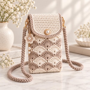 May include: A crochet crossbody phone bag featuring a cream and beige fan-shaped pattern. The bag has a flap closure with a single pearl button, a braided cord strap, and a dangling charm with a crochet flower and three small white pearls.