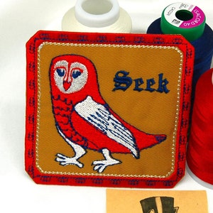 May include: An embroidered owl patch featuring a red and white owl with the word "Seek" on a brown fabric background. The patch is edged with a gold border and a red and white border.