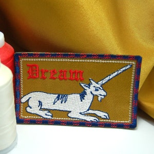 May include: A gold embroidered patch with a white unicorn and the word "Dream" in red lettering. The patch has a blue and red border.