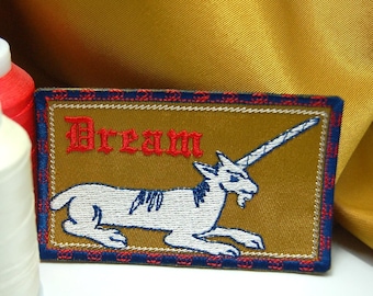 Op de afbeelding: Een gouden geborduurde patch met een wit eenhoorn en het woord "Dream" in rode letters. De patch heeft een blauwe en rode rand.