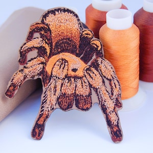 Peut inclure: Écusson brodé en forme d'araignée, dans des tons de marron et d'orange, avec des coutures détaillées. L'écusson est placé près de bobines de fil orange et marron, et d'un tissu beige plié.