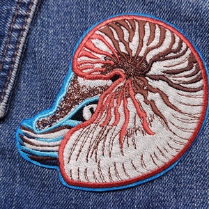 Puede incluir: Parche bordado con un diseño de concha de nautilus. El parche tiene un borde azul y muestra los intrincados detalles de la concha en tonos marrón, rojo y blanco. El parche está adherido a una chaqueta vaquera.