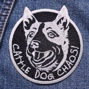 May include: A black and white embroidered patch with a dog's head in the center. The text "Cattle Dog Chaos!" is embroidered around the dog's head in white.