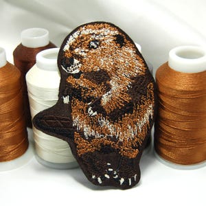 May include: Embroidered patch of a brown and white beaver with a black background. The patch is surrounded by spools of brown and white thread.