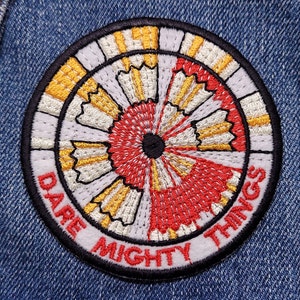 May include: A circular embroidered patch with a white, red, and yellow design. The text "Dare Mighty Things" is written around the edge of the patch.