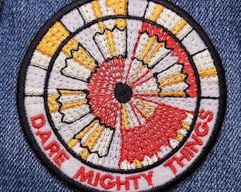 May include: A circular embroidered patch with a white, red, and yellow design. The text "Dare Mighty Things" is written around the edge of the patch.