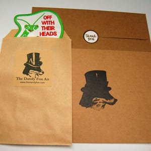 May include: Two brown paper envelopes with a green and white patch on the top envelope. The patch has the text "Off with their heads". The bottom envelope has a black and white illustration of a fox wearing a top hat. The text "The Dandy Fox Art" and a website address are printed on the top envelope.