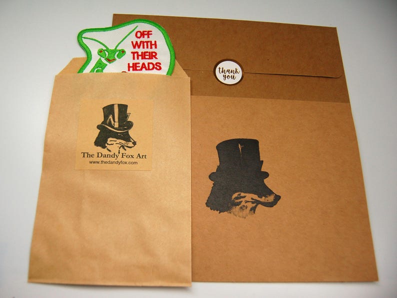 May include: Two brown paper envelopes with a green and white patch on the top envelope. The patch has the text "Off with their heads". The bottom envelope has a black and white illustration of a fox wearing a top hat. The text "The Dandy Fox Art" and a website address are printed on the top envelope.