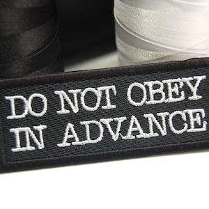 May include: Black embroidered patch with white text that reads "Do Not Obey In Advance".