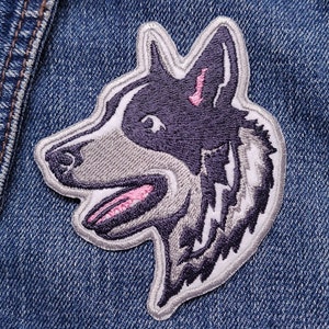 Op de afbeelding: Geborduurde strijkpatch van een grijs en zwart husky hondenhoofd met roze accenten. De hond kijkt naar rechts met zijn bek open.