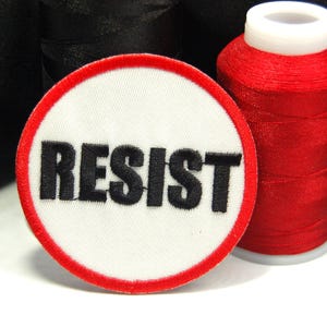 May include: A white embroidered patch with a red border and the word "RESIST" in black lettering.