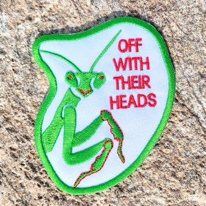 May include: A white patch with green embroidered edges and a green praying mantis with red eyes. The text "OFF WITH THEIR HEADS" is embroidered in red on the patch.