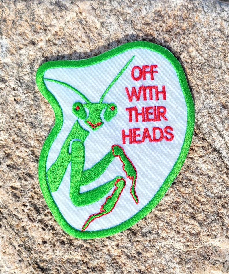 May include: A white patch with green embroidered edges and a green praying mantis with red eyes. The text "OFF WITH THEIR HEADS" is embroidered in red on the patch.