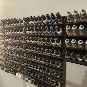 May include: A wall-mounted black tiered display rack holding numerous small plastic dropper bottles of acrylic paint. The bottles feature various colored caps and labels, organized in rows. A dark sculpted object is visible at the top edge.