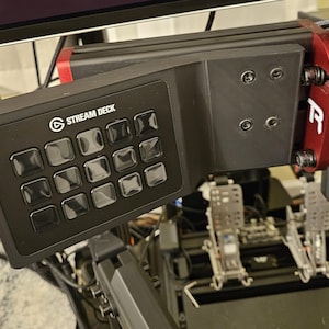 May include: A black Elgato Stream Deck with twelve square programmable buttons, mounted on a black and red metal racing simulator frame. The device features the white Stream Deck logo and is attached to the rig with visible black hex bolts.