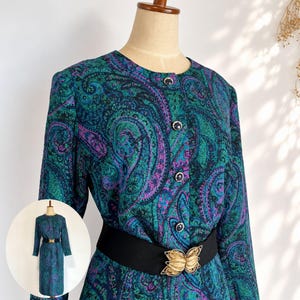 Pode incluir: Um vestido de manga comprida com um padrão paisley vibrante em tons de azul-petróleo, verde e roxo. O vestido tem um decote redondo, fecho frontal com botões escuros e um cinto elástico preto largo com uma fivela dourada em forma de borboleta.