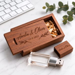 Op de afbeelding: Een rechthoekige houten doos met een schuifdeksel gegraveerd met de namen Amelia & Oliver, de datum 12-2-2023 en bladillustraties. De doos bevat houtsnippers en ligt naast een bijpassende houten USB-dop en een doorzichtige kristallen USB-stick.