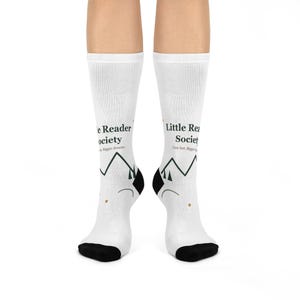 Könnte beinhalten: Ein Paar weiße Socken mit schwarzem Text, der Little Reader Society und Cozy feet. Bigger dreams. lautet. Das Design umfasst eine schwarze Berggrafik, einen kleinen goldenen Punkt sowie schwarze Zehen- und Fersenbereiche an jeder Socke.