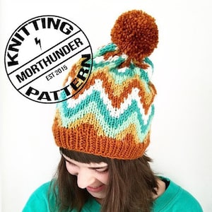 Puede incluir: Un gorro de punto con un patrón de chevron en tonos de naranja, amarillo, blanco y turquesa. El gorro tiene un pompón marrón en la parte superior. El gorro lo lleva puesto una persona con el pelo castaño.