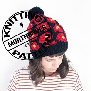 May include: Black knit beanie with a red and yellow floral pattern and a black pom pom on top. The beanie is being worn by a person with brown hair.