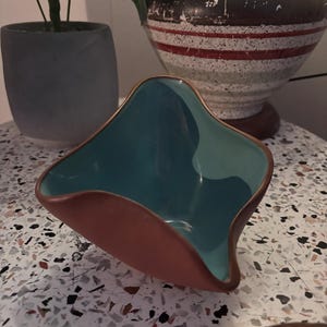 Może przedstawiać: Mała, nieregularnie ukształtowana ceramiczna miska z matową zewnętrzną stroną w kolorze terakoty oraz gładkim, błyszczącym wnętrzem w kolorze turkusowym. Miska posiada charakterystyczną, falistą krawędź, która wygina się do wewnątrz i na zewnątrz.