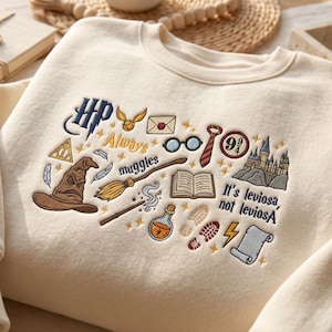 Może przedstawiać: Kremowa bluza z kolorowym haftem przedstawiającym różne magiczne ikony, w tym tiarę przydziału, miotłę, zamek oraz napisy "HP", "Always", "muggles" i "It's leviosa, not leviosA" umieszczone na przedniej części klatki piersiowej.