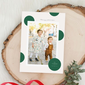 May include: A Christmas card with a photo of two children in front of a green background with white polka dots. The text "Merry Christmas" is written in cursive at the top of the card. The children are holding hands and smiling. The card also includes the text "The Tobins" in a circle.