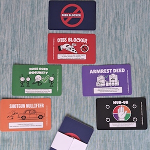 May include: A collection of rectangular game cards with various colors and illustrations. The cards feature text including DIBS BLOCKER, NOSE GOES IMMUNITY, ARMREST DEED, SHOTGUN NULLIFIER, and NUH-UH. One stack of cards is secured with a white paper band.