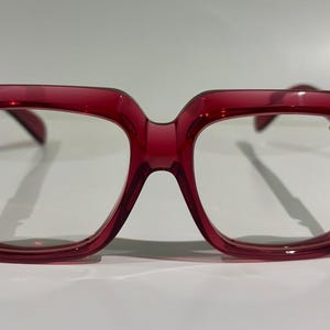 May include: Oversized square spectacles with a translucent deep red frame. The thick plastic rims feature silver-toned metal accents on the outer hinges. The brand name Cazal is printed in small gold lettering on the upper right lens area.