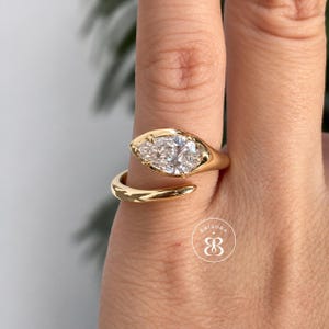 May include: A gold bypass ring featuring a pear-shaped white gemstone set in a polished yellow gold band. The open-ended design wraps around the finger, showcasing a clear, faceted stone held by prongs. A small circular logo with the text BRISORA is visible.