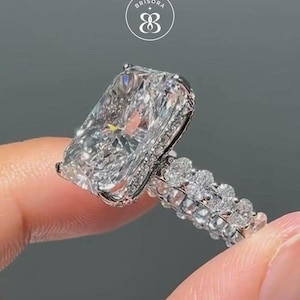 May include: A silver-toned ring featuring a large rectangular radiant-cut centre stone and a double band encrusted with small round stones. The ring displays the text BRISORA with a logo symbol above the centre stone on the grey background.
